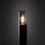 Lampa ogrodowa Ida H80 w kolorze czarnym z oprawą na żarówkę E27. Posiada szklany klosz o cylindrycznym kształcie, w którym widoczna jest stylizowana żarówka. Produkt stojący, ujęty z boku na jednolitym tle.