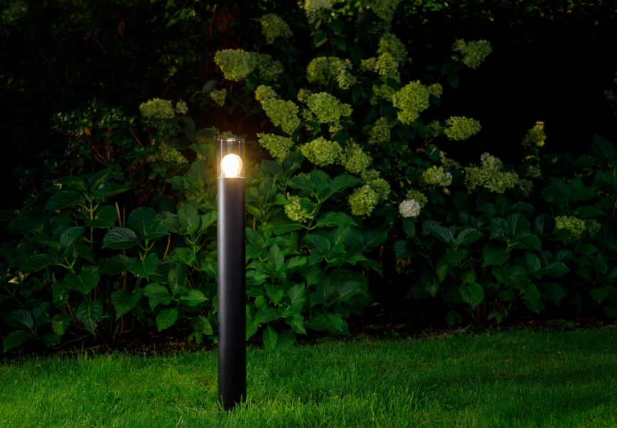 Czarna lampa ogrodowa Ida H80 z jedną oprawą E27, stojąca na trawniku. Posiada prostą, cylindryczną konstrukcję z przezroczystym kloszem, w którym widoczna jest żarówka. Tło to gęste, zielone krzewy i kwiaty.