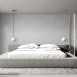 Złota lampa wisząca Dorado LP-002/1P z okrągłym, białym kloszem, zawieszona na długim kablu. Ujęcie frontalne, lampa umieszczona nad łóżkiem w minimalistycznej sypialni z szarymi ścianami i pościelą.