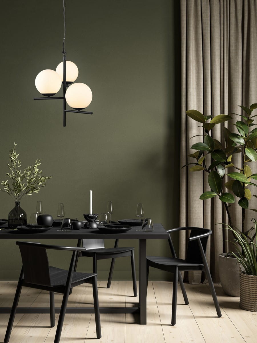 Lampa wisząca Pure w czarnym kolorze z trzema okrągłymi kloszami w jasnym odcieniu. Produkt zawieszony nad stołem w jadalni, otoczony ciemnymi krzesłami i dekoracjami, na tle zielonej ściany i zasłon.