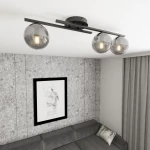 Kairo lampa sufitowa na trzy żarówki E14 w kolorze czarnym i grafitowym. Utrzymana w nowoczesnym stylu, z przezroczystymi, szarymi kloszami. Zainstalowana na suficie, w otoczeniu szarej ściany i kanapy.
