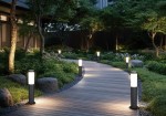 Lampa stojąca ogrodowa Trioto H45 w kolorze czarnym, z miejscem na jedną żarówkę E27. Ujęcie z boku, ukazujące lampę wśród zieleni, na drewnianym chodniku, z podświetleniem, które tworzy przytulną atmosferę.
