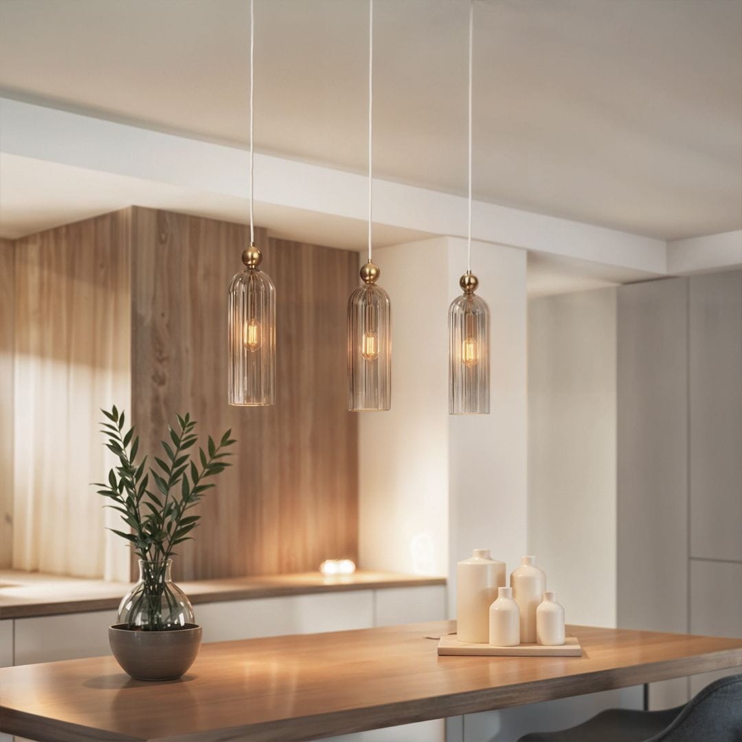 Lampa wisząca Regina w kolorze złotym z trzema kloszami w kształcie walca, wykonanymi z przezroczystego szkła. Zawieszona na białych przewodach, umieszczona nad drewnianym blatem, obok dekoracyjnej rośliny w wazonie.