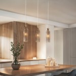 Lampa wisząca Regina w kolorze złotym z trzema kloszami w kształcie walca, wykonanymi z przezroczystego szkła. Zawieszona na białych przewodach, umieszczona nad drewnianym blatem, obok dekoracyjnej rośliny w wazonie.