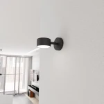 Kinkiet Sora 1xGX53 w kolorze czarnym zamontowany na ścianie. Posiada cylindryczny klosz i okrągłą podstawę. W tle widać nowoczesne wnętrze z dużymi oknami i minimalistycznym wystrojem.