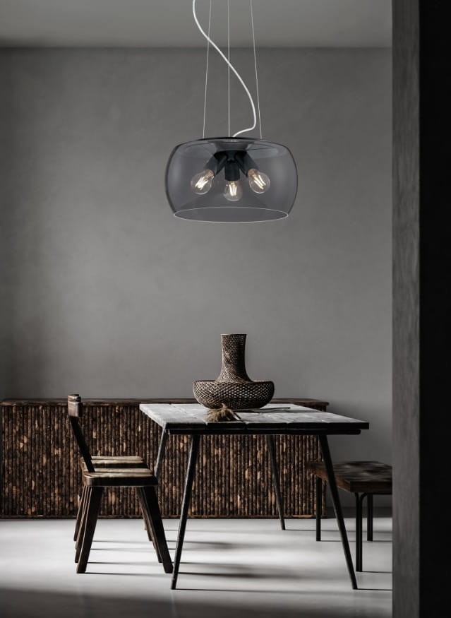 Lampa wisząca Valente w kolorze antracytowym z trzema oprawkami E27, z przezroczystym kloszem, zawieszona na cienkich linkach. W tle drewniane meble i stół, tworzące nowoczesną aranżację wnętrza.
