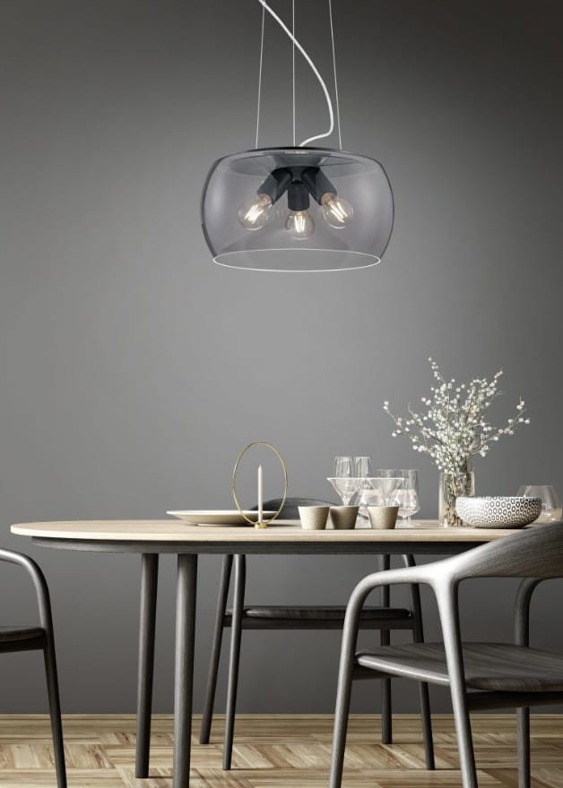 Lampa wisząca Valente w kolorze antracytowym z trzema źródłami światła E27, umieszczona nad okrągłym drewnianym stołem. Przezroczysta osłona podkreśla elegancki design. W tle szare ściany oraz dekoracje stołowe, takie jak kwiaty i naczynia.