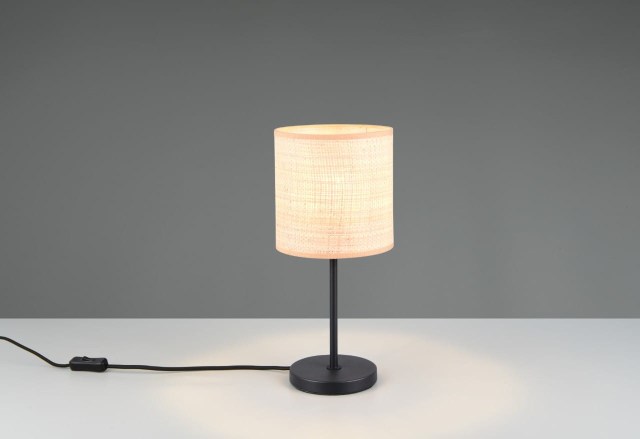 Lampa stołowa Aimee z czarną podstawą i naturalnym kloszem, stojąca na okrągłej podstawie. Klosz ma cylindryczny kształt, wytworzony z materiału, emitujący ciepłe światło. Kabel z włącznikiem widoczny obok podstawy. Tło szare, proste.