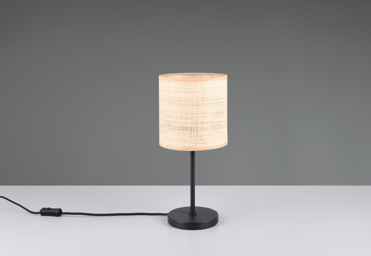 Lampa stołowa Aimee z czarną podstawą i naturalnym, teksturowanym kloszem z plecionki. Ujęcie frontalne, lampa stoi na białym stole, z widocznym kablem z włącznikiem. Tło w kolorze szarym.