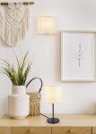 Lampa stołowa Aimee 1xE27 w kolorze czarnym z naturalnym abażurem. Ustawiona na drewnianym stoliku obok jasnego wazonu z trawami dekoracyjnymi i wiklinowym koszykiem, w tle obraz z roślinnym motywem.
