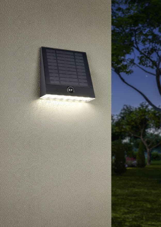 Kinkiet zewnętrzny Alvorada Hybrid w kolorze czarnym, wyposażony w panel solarny i 1xLED o mocy 6W. Zamontowany na ścianie, emitujący światło, z widocznym czujnikiem. Tło przedstawia zewnętrzną przestrzeń z roślinnością.
