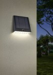 Kinkiet zewnętrzny Alvorada Hybrid w kolorze czarnym, wyposażony w panel solarny i 1xLED o mocy 6W. Zamontowany na ścianie, emitujący światło, z widocznym czujnikiem. Tło przedstawia zewnętrzną przestrzeń z roślinnością.