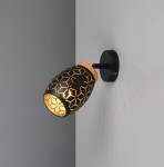 Kinkiet Bidar z czarnym kloszem w geometryczny wzór, zamontowany na ścianie. Klasyczny, regulowany punkt świetlny z oprawką na żarówkę E14, z drewnianym wykończeniem u góry. Oświetlenie emituje ciepłe światło, tworząc przytulną atmosferę.