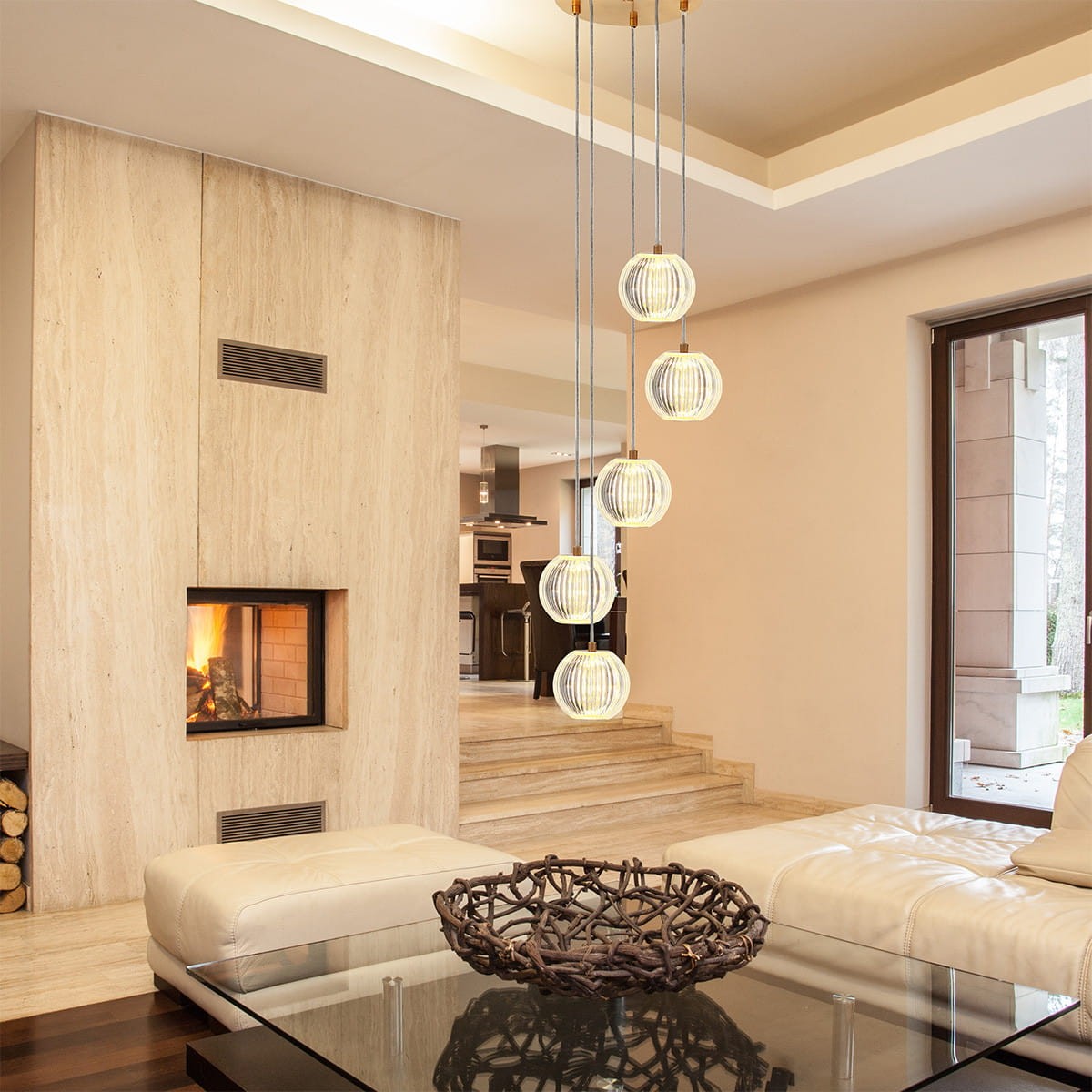 Lampa wisząca Brillant z pięcioma kloszami LED w kolorze złotym, zawieszona na cienkich linkach. Klosze mają okrągły kształt z transparentnym wzorem. Lampa znajduje się w jasnym, nowoczesnym wnętrzu z kominkiem i sofą.