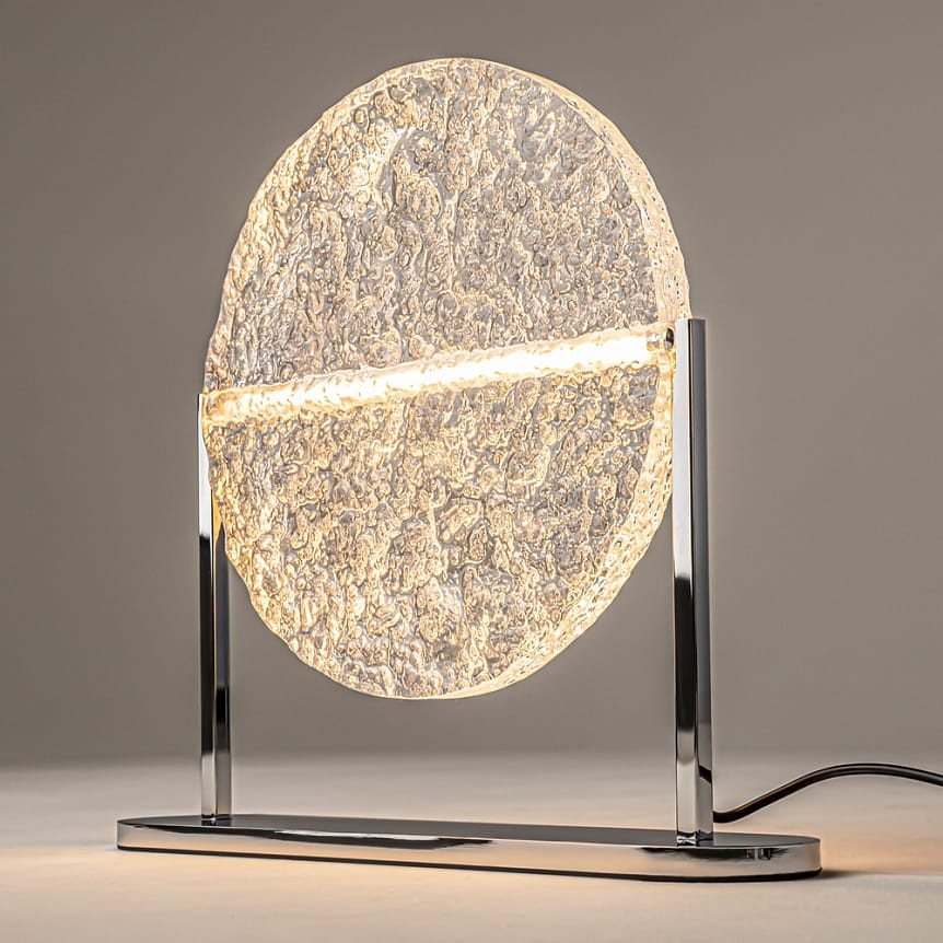 Lampa stołowa LED Mangata o mocy 7W w kolorze chromu. Posiada okrągły klosz z teksturowanego szkła, zamontowany na metalowej podstawie. Ujęcie frontalne, lampa stoi na płaskiej powierzchni, emitując ciepłe światło.