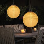 Lampa wisząca solarna Origami D20 w kształcie okrągłego lampionu, w kolorze białym, z wyraźnymi, poziomymi fałdami. Zawieszona nad drewnianym stołem, w otoczeniu świec, idealna do ogrodu. Świeci ciepłym światłem, tworząc przytulną atmosferę.