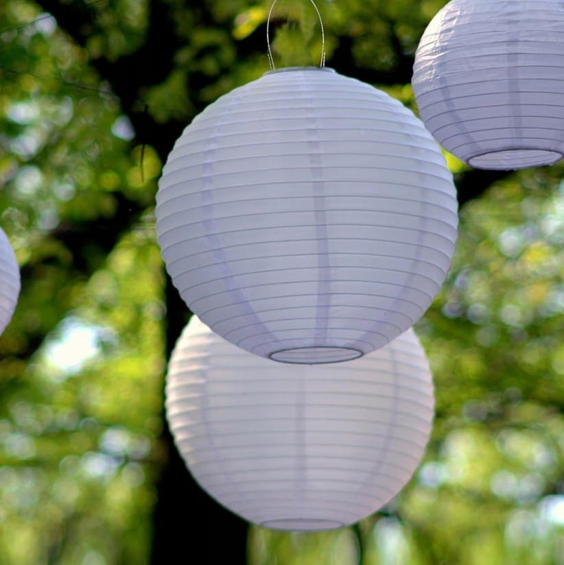 Lampa wisząca solarna Origami D20 w kształcie białej, okrągłej latarni z delikatnymi, poziomymi żłobieniami. Zawieszona w zielonym otoczeniu, tworzy nastrojową atmosferę. Widoczne elementy montażowe na górze.