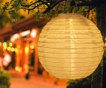 Biała lampa wisząca solarna Origami D20 w kształcie okrągłej latarni, zawieszona na gałęzi drzewa. Wykonana z materiału o delikatnej teksturze, emituje ciepłe światło w otoczeniu zieleni i rozmytych świateł.