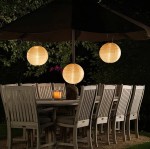 Lampa wisząca solarna Origami D20 w kształcie białej kuli, zawieszona pod parasolem, oświetla drewniany stół w ogrodzie. W tle widać kilka drewnianych krzeseł oraz roślinność. Świeca w szklanym pojemniku na stole dodaje przytulnego klimatu.