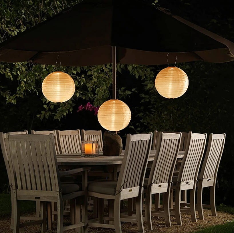 Lampa wisząca solarna LED Origami D30 w białym kolorze, zawieszona pod parasolem. W tle stół z drewnianymi krzesłami, otoczenie nocne, z delikatnym oświetleniem oraz roślinnością. Widać ciepłe światło lampy oraz świecę na stole.