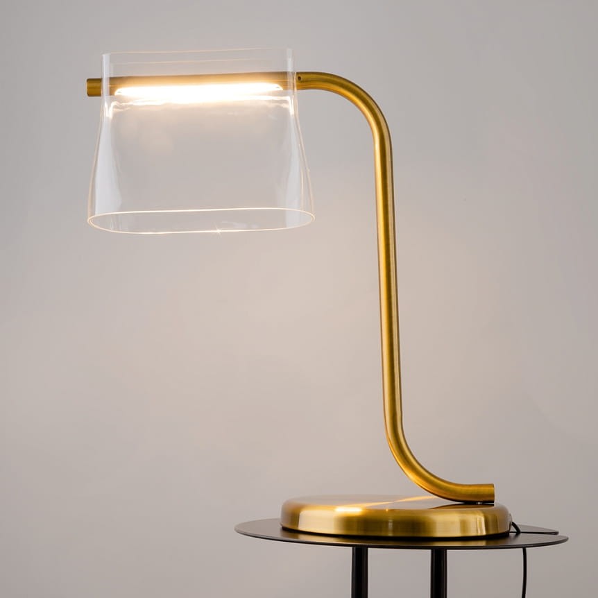 Lampa stołowa LED Cabaret o mocy 8W, z mosiężnym stelażem i przezroczystym kloszem. Ujęcie frontalne, lampa ustawiona na okrągłej podstawie. Delikatne światło emitowane z górnej części klosza w kształcie stożka. Tło jasne, neutralne.