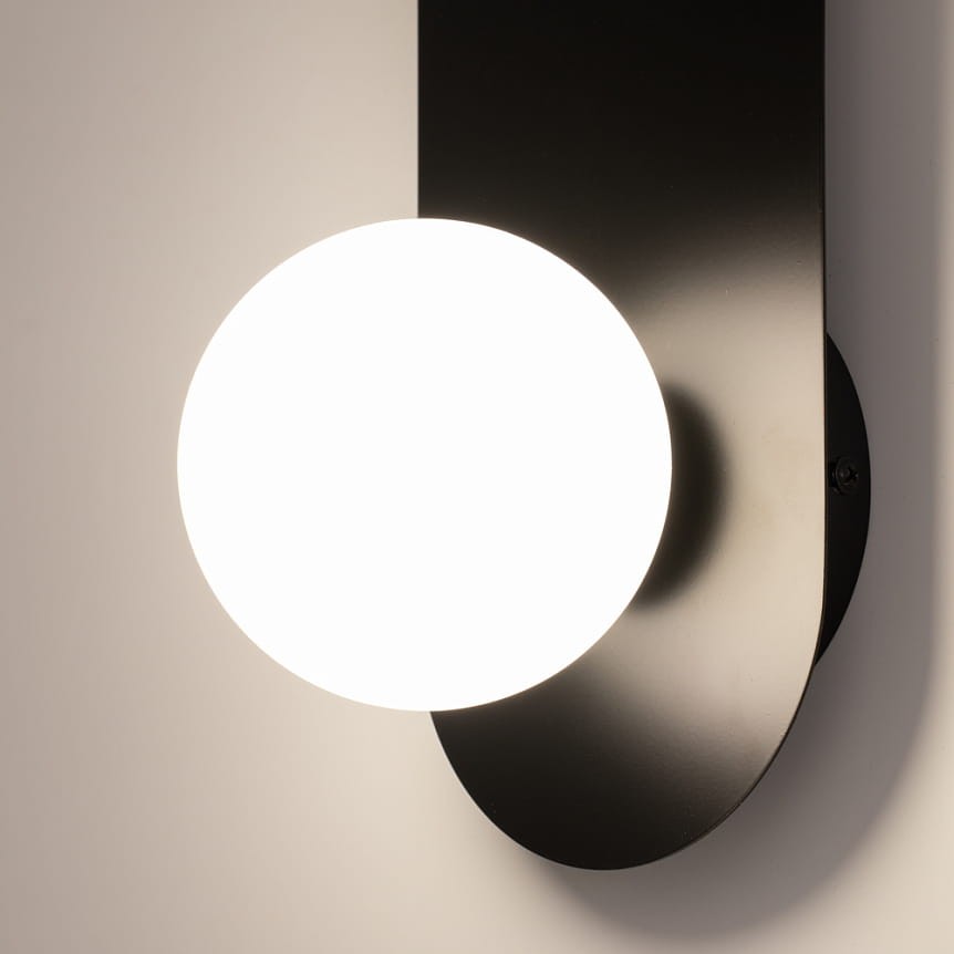 Kinkiet Bao 1xG9 w kolorze czarnym, z okrągłą, białą żarówką. Montaż ścienny, widoczny z boku. Wykonanie z matowego metalu, minimalistyczny design, idealny do nowoczesnych wnętrz. Tło jasne, podkreślające formę lampy.