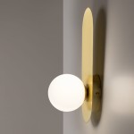 Kinkiet Bao MOD419WL-01G w kolorze złotym, zamontowany na ścianie, z okrągłą, matową, białą żarówką oraz prostokątną, gładką, złotą podstawą. Produkt widoczny na tle szarej ściany.