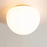 Lampa sufitowa Glassy z kulistym kloszem w kolorze białym i złotą podstawą. Ujęcie z bliska pokazujące gładką, matową powierzchnię klosza. Lampa zamontowana na suficie, emanująca miękkie światło. Idealna do nowoczesnych wnętrz.