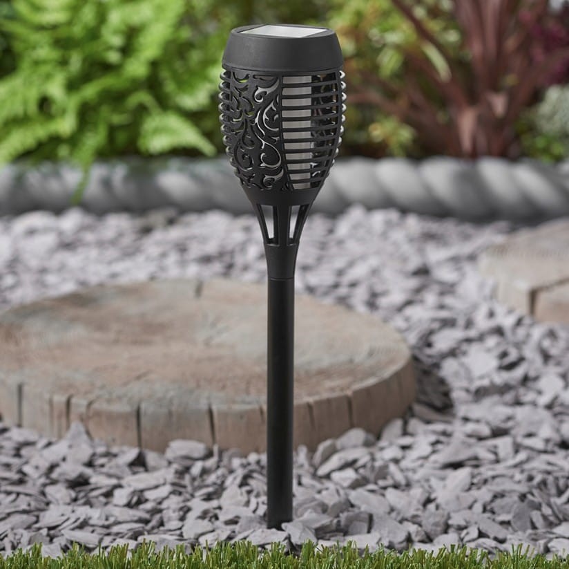 Czarna lampa solarna ogrodowa typu pochodnia, wbijana, z dekoracyjną obudową i LED, stojąca na tle kamieni i trawy, z widocznym wzorem na korpusie. Wysokość 50 cm, idealna do oświetlenia przestrzeni na zewnątrz.