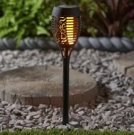 Czarna lampa solarna ogrodowa w kształcie pochodni, z dekoracyjnym wzorem, wbijana w ziemię. Wyposażona w diodę LED, emituje ciepłe światło. Ujęcie z boku, na tle żwirku i otaczających roślin. Wysokość 50 cm.