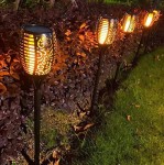 Czarna lampa solarna ogrodowa w postaci pochodni, w zestawie 4 sztuki, z dekoracyjnym wzorem. Stojąca, wbijana w ziemię, emituje ciepłe światło LED. Widoczna na tle ciemnych liści i zielonej trawy.