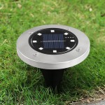 Lampa solarna ogrodowa Terra wbijana, srebrna, z okrągłą podstawą. Widoczne panele słoneczne i diody LED, umieszczona na trawie. Produkt w pozycji stojącej, przeznaczony do oświetlenia ogrodu lub ścieżek.