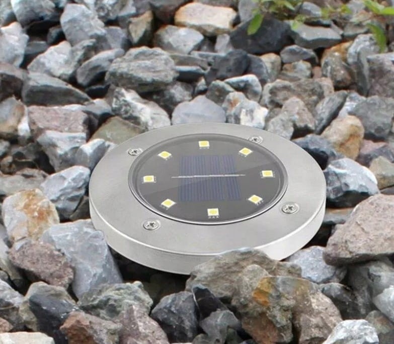 Lampa solarna Terra wbijana, srebrna, z okrągłą obudową, umieszczona na kamieniach. Widoczny panel słoneczny oraz sześć diod LED. Produkt leży płasko, na jednolitym tle z szaro-brązowych kamieni.