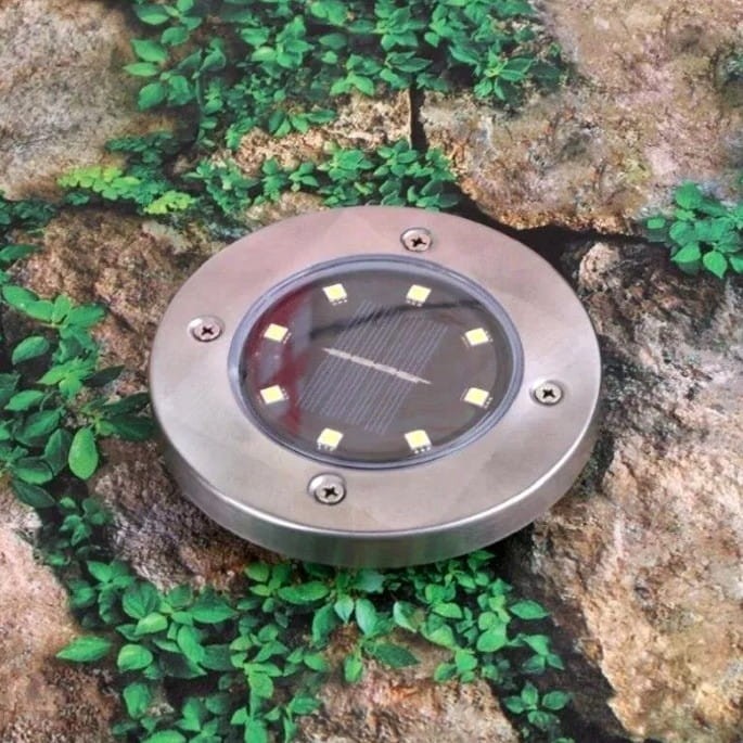 Lampa solarna ogrodowa wbijana Terra w srebrnym kolorze, okrągła, z przezroczystą soczewką, ośmioma diodami LED oraz widocznymi śrubami mocującymi. Ujęcie z góry na tle zielonych liści i kamieni, pokazujące szczegóły wykonania.