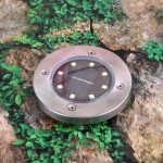 Lampa solarna ogrodowa wbijana Terra w srebrnym kolorze, okrągła, z przezroczystą soczewką, ośmioma diodami LED oraz widocznymi śrubami mocującymi. Ujęcie z góry na tle zielonych liści i kamieni, pokazujące szczegóły wykonania.