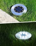 Lampa solarna ogrodowa Terra w kolorze srebrnym, wbijana w ziemię. Widoczna na trawie, z panelami słonecznymi i sześcioma diodami LED. Ujęcie z boku, ukazujące zarówno wyłączoną, jak i włączoną lampę emitującą światło.