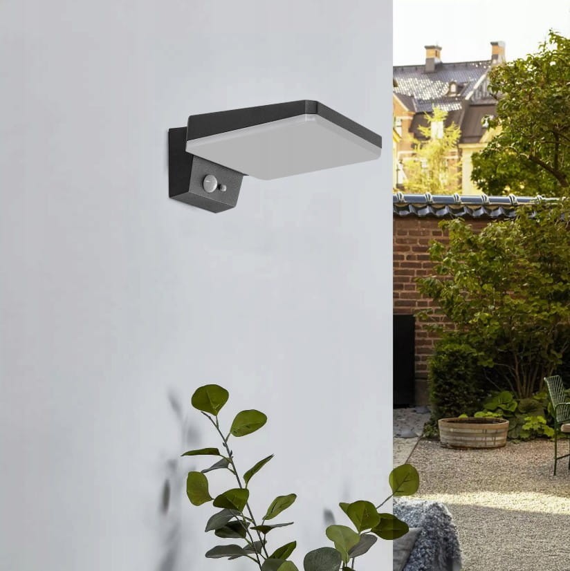 Kinkiet elewacyjny solarny Sonda S LED w kolorze czarnym, zamontowany na ścianie. Posiada czujnik ruchu i zmierzchu. W tle widoczna zieleń oraz elementy aranżacji ogrodowej.