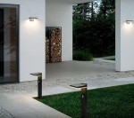 Kinkiet elewacyjny Sonda S LED w kolorze czarnym, z czujnikiem ruchu i zmierzchu, zamontowany na ścianie budynku. W tle widoczne drewno w przechowalni, a na trawniku stoją słupki solarne. Oświetlenie podkreśla nowoczesny design przestrzeni zewnętrznej.