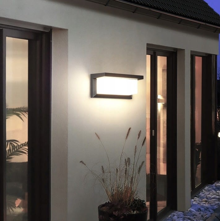 Czarny kinkiet elewacyjny Vasco LED z czujnikiem ruchu i zmierzchu, zamontowany na ścianie budynku. Oświetlenie emituje miękkie światło, otoczone przez roślinność w donicy na ziemi, w tle widoczne okna.