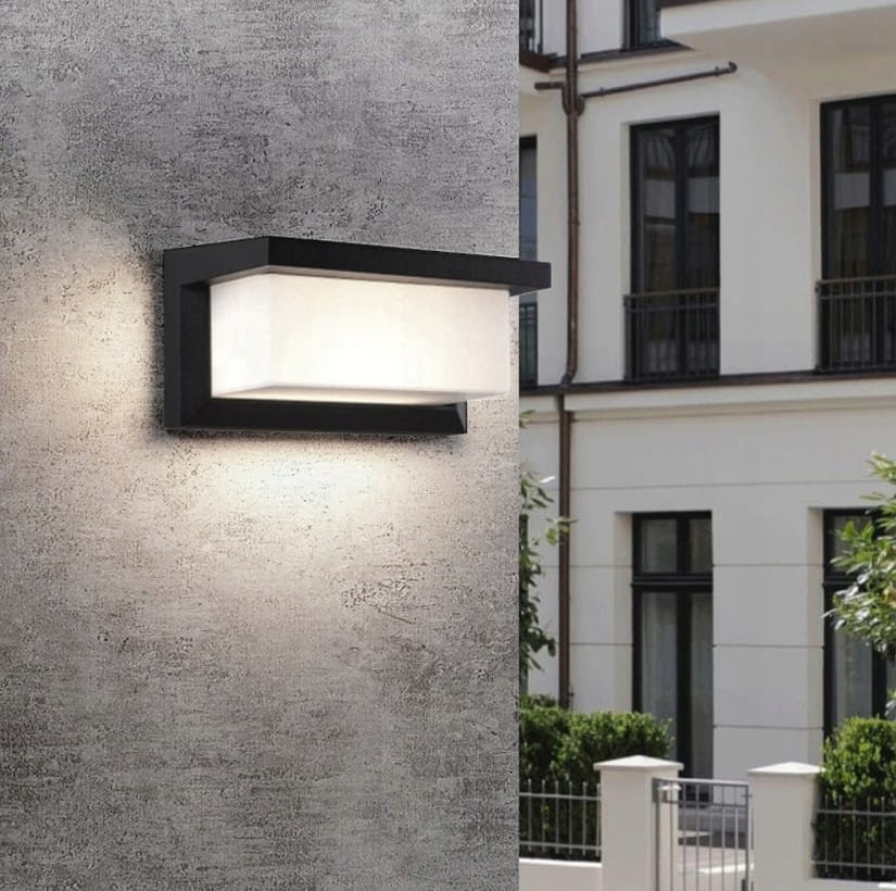 Vasco LED kinkiet elewacyjny solarny w kolorze czarnym z czujnikiem ruchu i zmierzchu, zamontowany na szarej ścianie. Oświetlenie ma prostokątny kształt, z przezroczystą osłoną, emitującym jasne światło w otoczeniu zieleni i budynków.