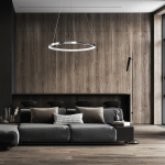 Lampa wisząca LED o mocy 27W w chromowanej obudowie, w kształcie okręgu. Zawieszona na cienkim przewodzie w nowoczesnym wnętrzu z drewnianą ścianą, nad ciemnoszarym narożnikiem z poduszkami. W tle widoczne czarne krzesła barowe.