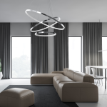 Lampa wisząca Hoop z trzema okręgami LED o chromowanej powierzchni, zawieszona w nowoczesnym wnętrzu. Widoczna w kontekście salonu z szarymi zasłonami, jasnymi oknami oraz minimalistycznym umeblowaniem w neutralnych kolorach.
