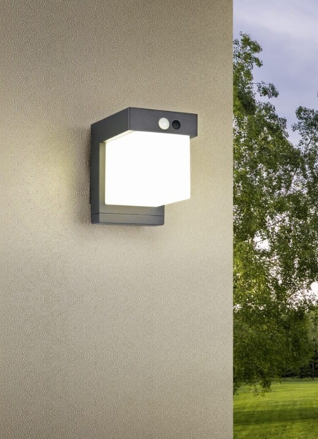 Kinkiet zewnętrzny Colombo Hybrid w kolorze czarnym, z białą, prostokątną kloszem LED 5,5W. Montowany na ścianie, z czujnikiem. Ochrona IP44, idealny do użytku na zewnątrz. Tło w neutralnym kolorze, z widoczną zielenią w tle.