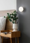 Kinkiet Earl z białą oprawą LED o mocy 1,5W, zamontowany na szarej ścianie obok łóżka. W tle widoczna drewniana rama łóżka, stół nocny z bambusa i doniczka z rośliną.