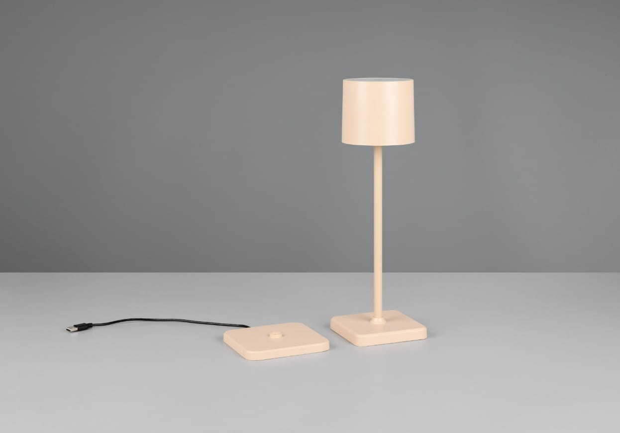 Lampa stołowa zewnętrzna Fernandez w kolorze piaskowym, z cylindrycznym kloszem i prostą podstawą. Ujęcie frontalne, lampa stoi na stole obok kwadratowej podstawy z przewodem USB. Wysokość lampy pozwala na oświetlenie małych przestrzeni.