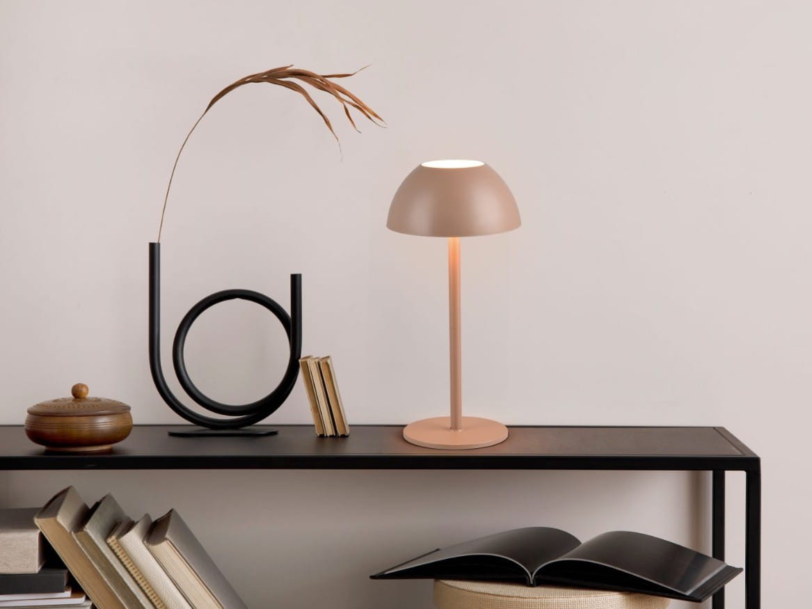 Lampa stołowa Ricardo w kolorze kawowym, z jedną żarówką LED 1,7W. Posiada zaokrąglony klosz oraz smukłą podstawę. Ustawiona na czarnej półce obok książek i dekoracji, w prostym, jasnym tle.