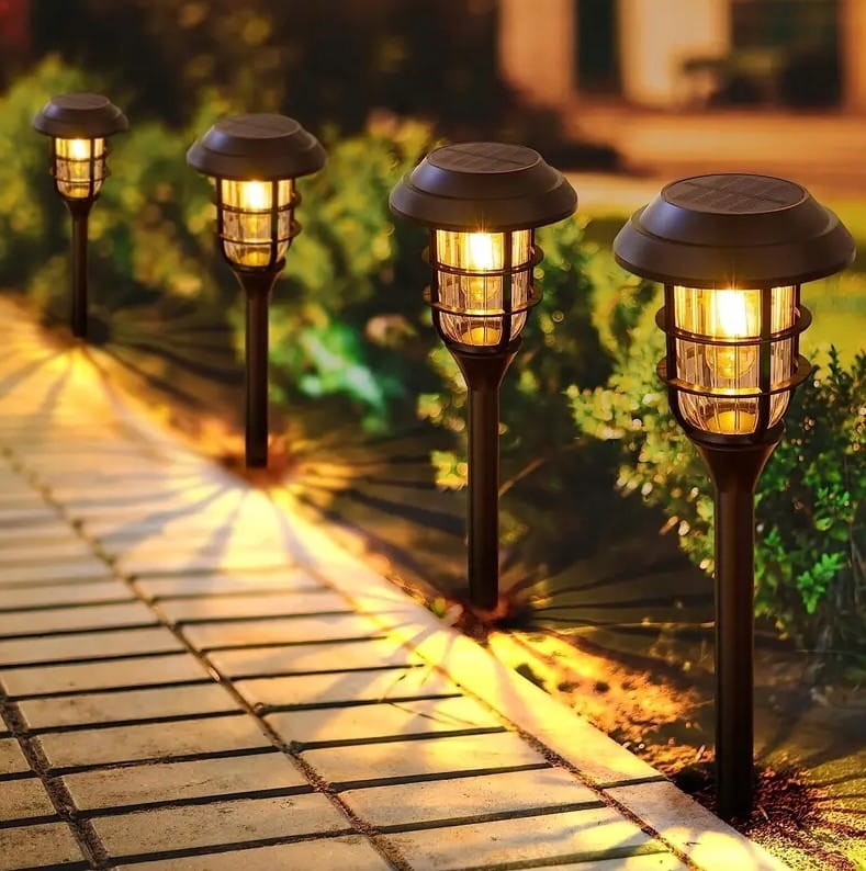 Latarnia solarna ogrodowa, zestaw 6 sztuk, czarna, wysoka na 45 cm. Oświetlenie LED z kloszem w metalowej ramie, wbijana w ziemię. Ujęcie z boku, na tle zieleni i chodnika, z włączonym światłem, rzucającym cienie na nawierzchnię.