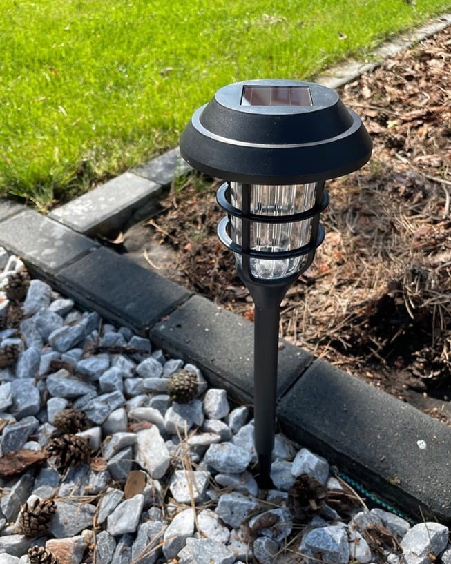 Latarnia solarna ogrodowa, czarna, wbijana, z przezroczystym kloszem, wysokość 45 cm, LED Premium. Ujęcie z boku, umieszczona na podłożu z kamieni, w tle trawa. Widoczne panele słoneczne na górze lampy.