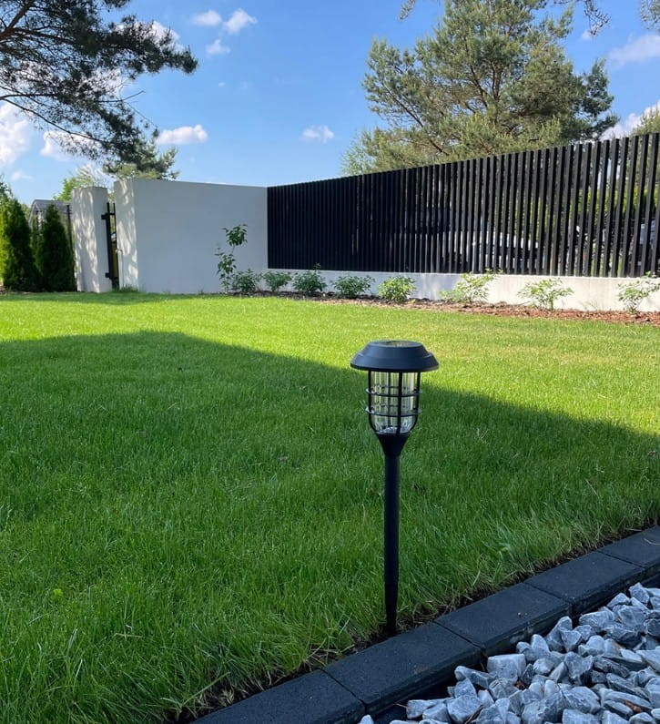 Latarnia solarna ogrodowa w kolorze czarnym, wbijana w ziemię, z lampą LED. Ujęcie frontalne, stojąca na trawniku, w tle biała ściana i ogrodzenie. Dobre oświetlenie, widoczne detale siatki w kloszu.
