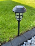 Lampa solarna ogrodowa Latarnia II w kolorze czarnym, wbijana, o wysokości 45 cm. Posiada przezroczysty klosz z metalową siatką. Ukazana w ogrodzie na tle zielonej trawy i szarej kostki brukowej.